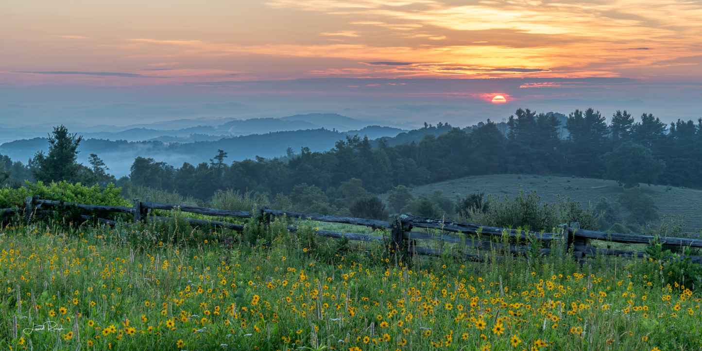 A scenic landscape photograph of a sunset over rolling hills and distant mountains, with a wooden fence and a field of yellow wildflowers in the foreground. 1x2 crop