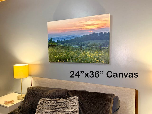 A 24x36 canvas wall art print depicting a scenic sunset over a green valley with yellow wildflowers, displayed on a bedroom wall above a bed with a text overlay indicating the size.