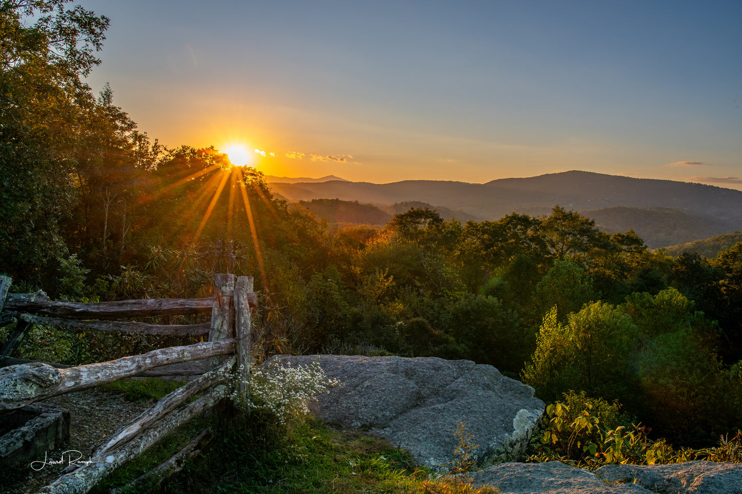View of sunset from Raven Rock overlook on the blue ridge parkway near  blowing rock, North Carolina, with a fence, wildflowers and large rocks in the foreground.