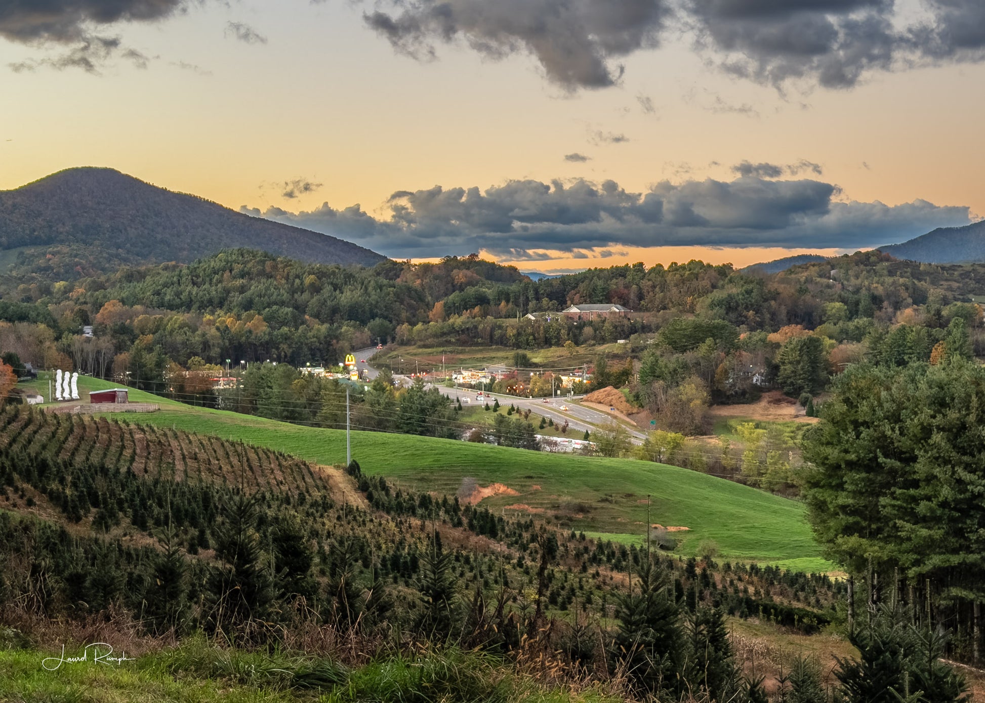 Scenic view of West Jefferson, North Carolina crossroads (221 and Jefferson Ave) with trees and mountains under a dramatic sky at sunset.