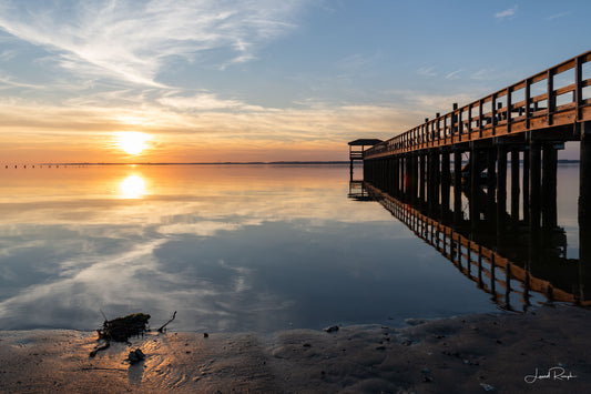 Fort Fisher pier on the Cape Fear River in North Carolina at sunset with calm, glassy water reflecting the pastel sky.