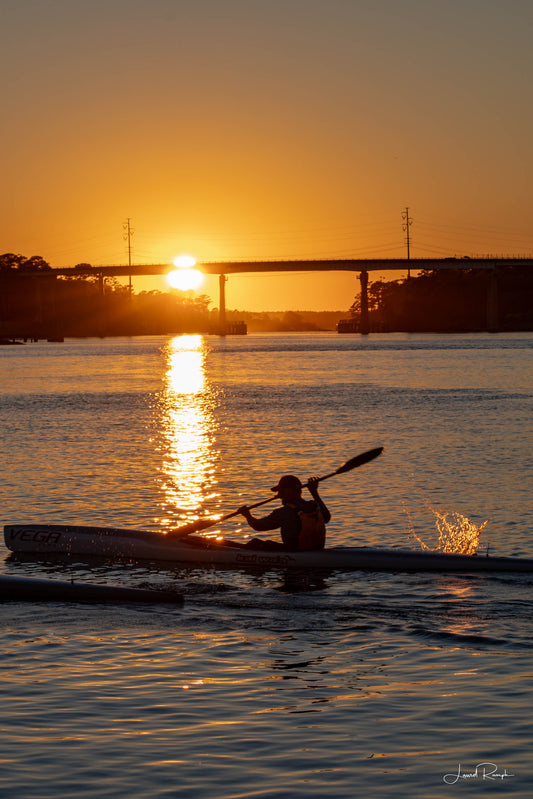 Kayaker at Snow’s Cut Marina, North Carolina with sunset behind Snow’s Cut Bridge and water splashing from the paddle.