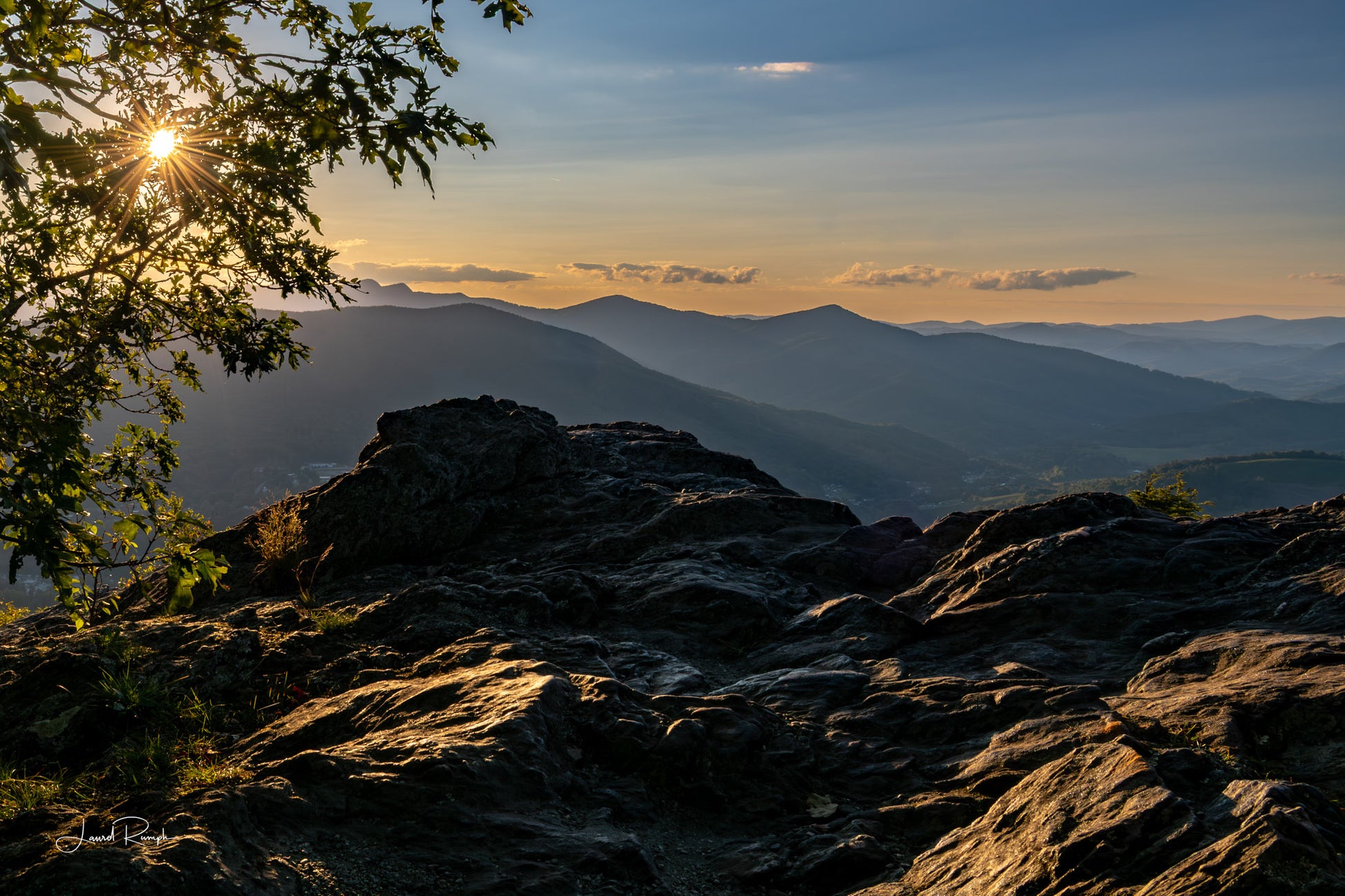 Sunset view from Jefferson Overlook on top of Mount Jefferson in Ashe County, North Carolina, with a sun starburst peeking from the tree and large rock formation in the foreground. 2x3 crop