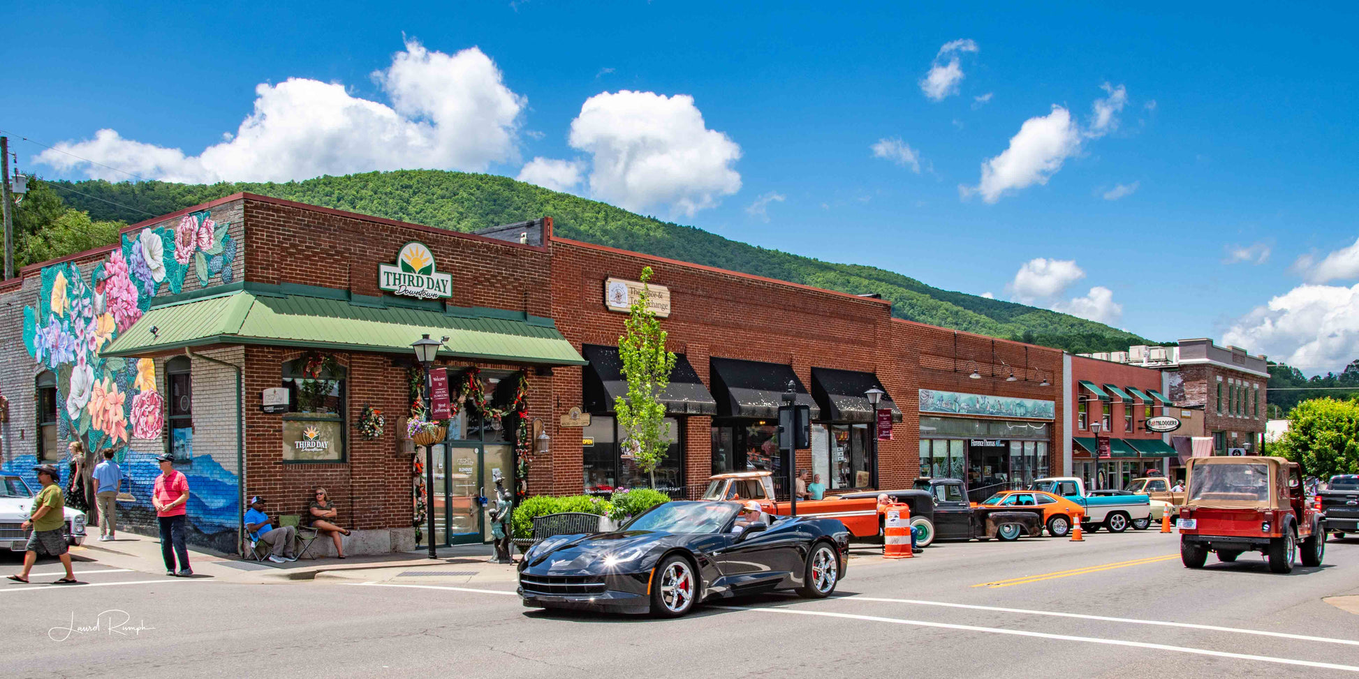 Cruise-In event in West Jefferson, North Carolina with a Corvette on a small-town street, shops and people nearby, and mountains in the distance.